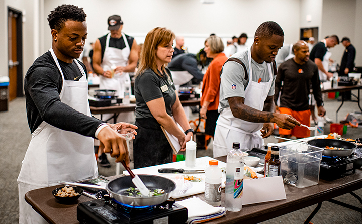 Rookie cornerback Shaun Jolly (l) works on his dish while BW catering manager Jennie Vassanelli gets a sneak peek ahead of judging.