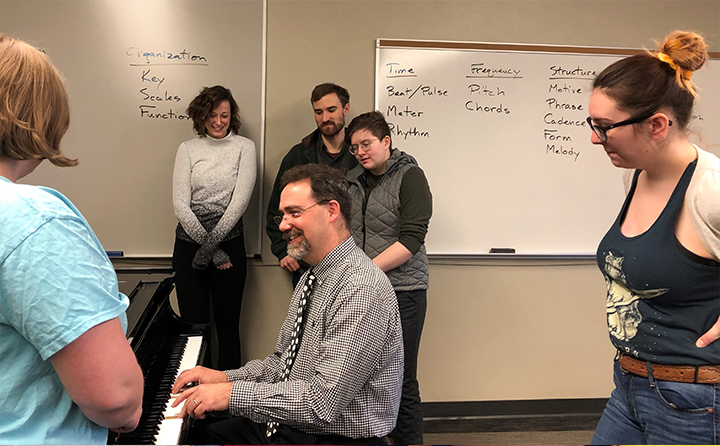 Honors students standing around BW professor playing piano in classroom