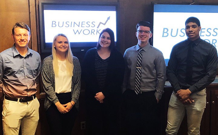 Photo of students standing in a conference room in front of TV screens with the Business Works logo